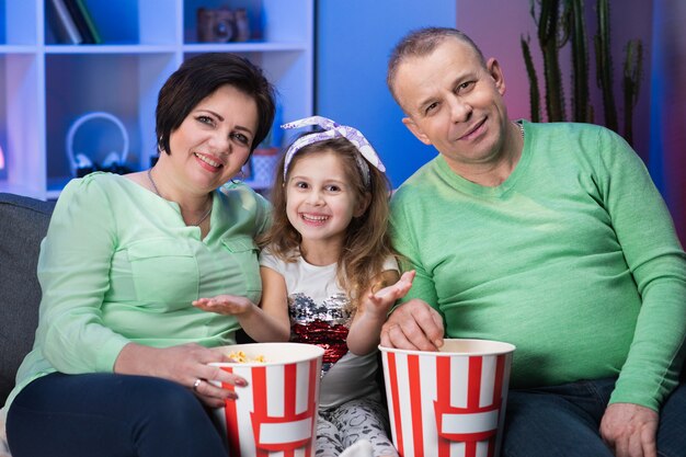 Lächelnde Großeltern und Enkelin mit Enkelin, die zu Hause auf dem Sofa sitzt. Großvater und Großmutter mit Enkelin sitzen auf Sofa zu Hause und schauen sich einen Film an.