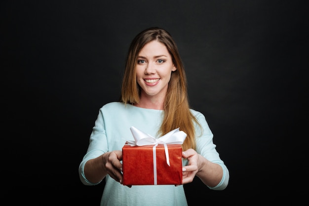 Lächelnde großzügige junge Frau, die Freude ausdrückt und Geschenkbox gibt, während sie gegen schwarze Wand steht