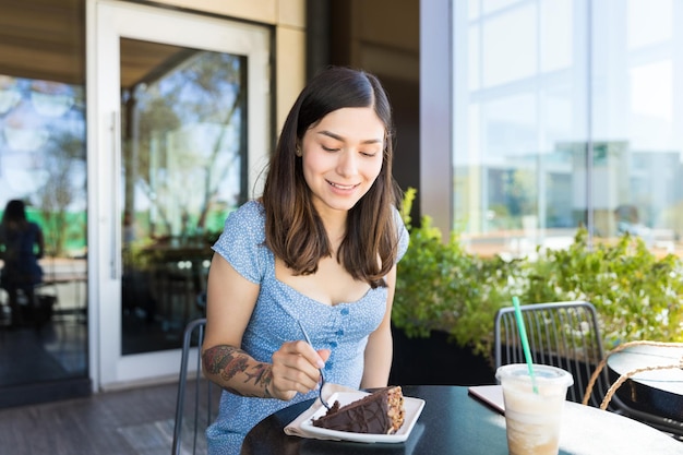 Lächelnde hispanische Brünette, die ein Stück Kuchen isst, während sie im Café sitzt