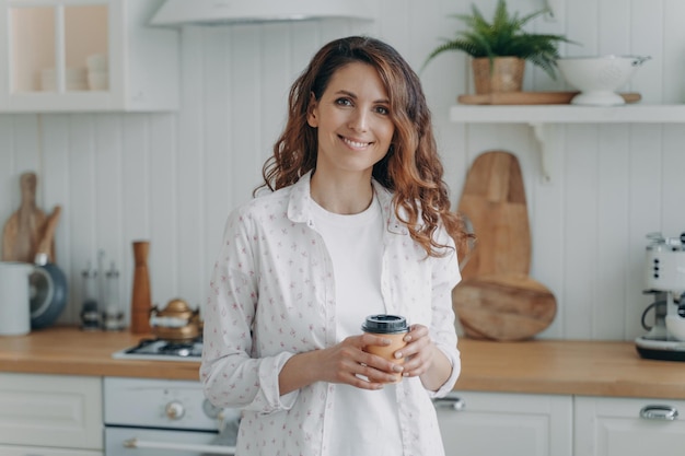 Lächelnde hispanische Frau mit einer Papptasse Kaffee, die in einer gemütlichen, modernen Küche steht und in die Kamera blickt