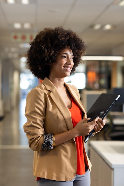 Lächelnde hispanische Geschäftsfrau mit Afro-Frisur hält Smartphone und Laptop am Arbeitsplatz