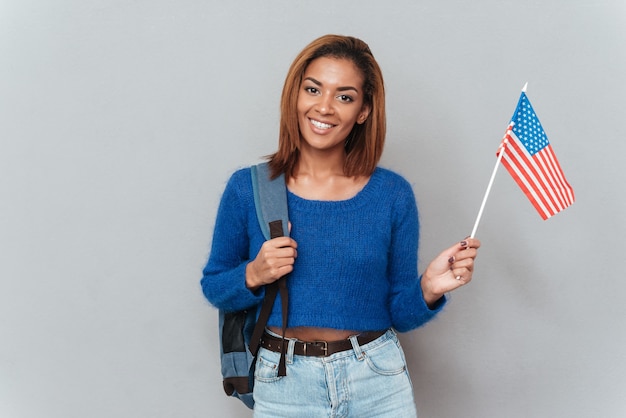 Lächelnde hübsche afrikanische Frau in Pullover und Jeans mit Rucksack und mit Flagge der USA. Isolierter grauer Hintergrund