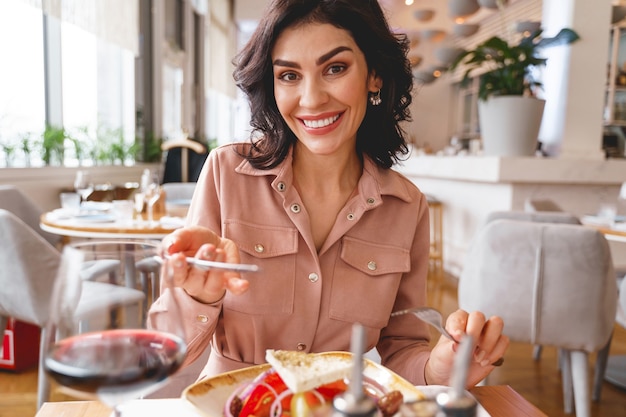 Lächelnde hübsche Dame mit Gabel und Messer beim Sitzen am Tisch mit leckerem, gesundem Essen und Wein