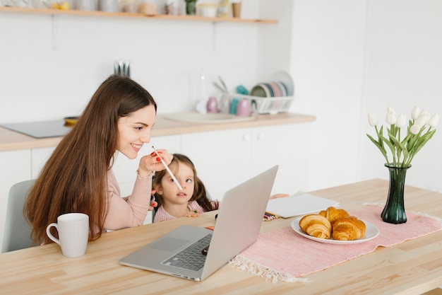 Lächelnde hübsche Frau beim Schminken, während sie mit ihrer kleinen Tochter in der Küche während des Frühstücks sitzt.