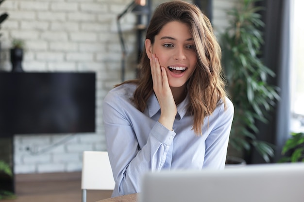 Lächelnde hübsche Frau, die am Tisch sitzt und Laptop-Bildschirm betrachtet. Glücklicher Unternehmer, der Nachrichten-E-Mails mit guten Nachrichten liest und online mit Kunden chattet.