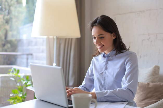 Lächelnde hübsche Frau, die am Tisch sitzt und Laptop-Bildschirm betrachtet. Glücklicher Unternehmer, der Nachrichten-E-Mails mit guten Nachrichten liest und online mit Kunden chattet.