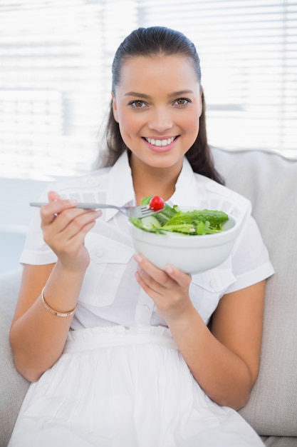 Lächelnde hübsche Frau, die den gesunden Salat sitzt auf Sofa isst
