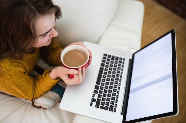 Lächelnde hübsche Frau, die Laptop beim Trinken des Kaffees auf Couch im Wohnzimmer verwendet