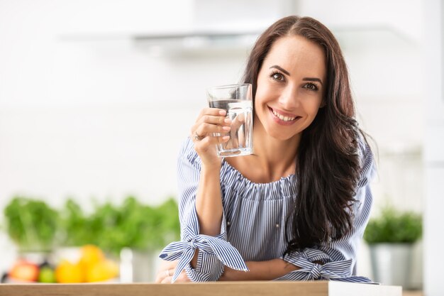 Lächelnde hübsche Frau hält ein Glas Wasser, das sich auf den Küchentisch stützt.