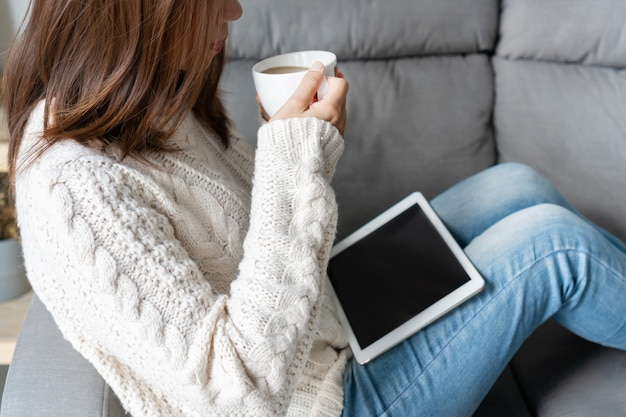 Lächelnde hübsche Frau im weißen Pullover, der Kaffee trinkt, während sie auf einem Sofa im Wohnzimmer zu Hause sitzt