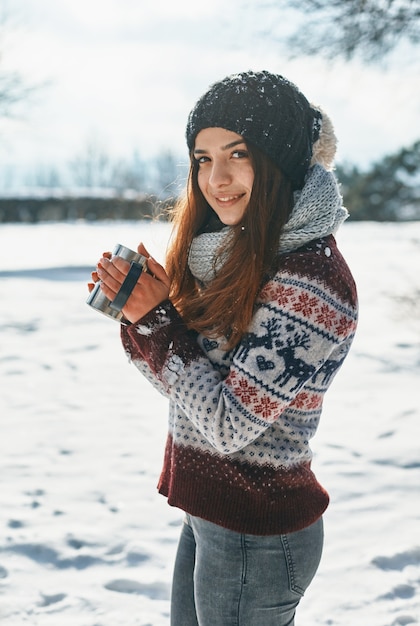 Lächelnde hübsche Frau mit Tasse heißem Getränk, die nahe Winterwald steht und Ferien unter atemberaubender Winterlandschaft verbringt