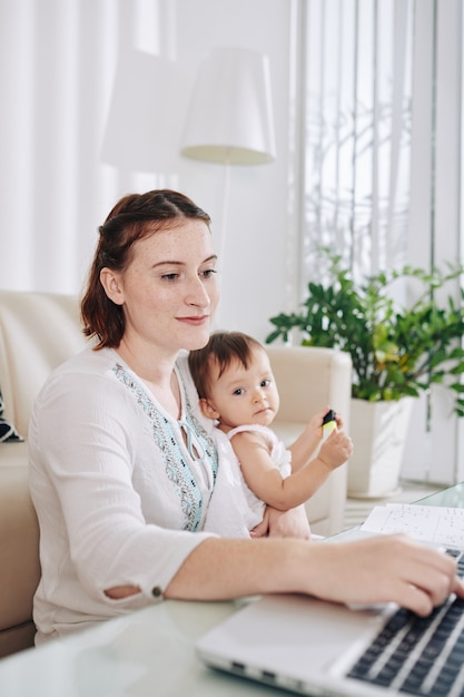 Lächelnde hübsche junge Frau, die aufgrund einer Pandemie zu Hause bleibt und mit Baby auf dem Schoß am Laptop arbeitet