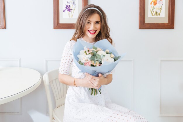 Lächelnde hübsche junge Frau im weißen Kleid, die im Café sitzt und Blumenstrauß hält