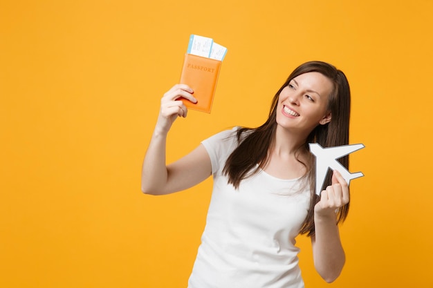 Lächelnde hübsche junge Frau in weißer Freizeitkleidung mit Reisepass, Bordkarten, Papierflugzeug isoliert auf gelb-orangefarbenem Hintergrund im Studio. People-Lifestyle-Konzept. Mock-up-Kopienbereich.