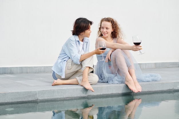 Lächelnde hübsche junge Frauen, die am Schwimmbad sitzen, Wein trinken und Ideen diskutieren