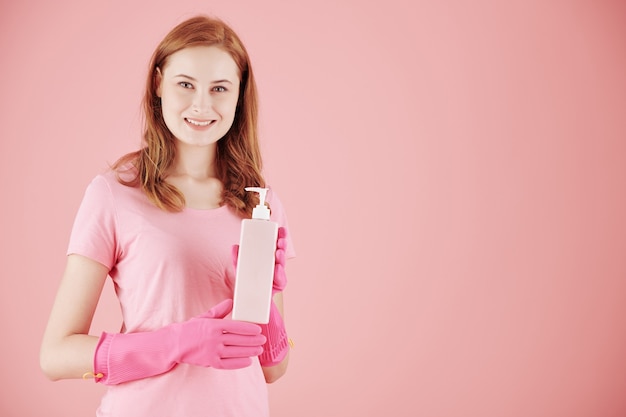 Lächelnde hübsche junge Hausfrau in rosa Gummihandschuhen, die Flasche Reinigungsmittel zeigen