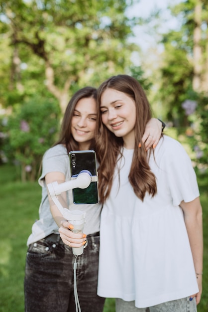 Lächelnde hübsche junge Influencer-Bloggerinnen filmen oder nehmen Videos mit ihrem Handy auf einem Stabilisator in einem sonnigen grünen Park draußen auf. Blogging-Konzept. Weicher selektiver Fokus, Defokussierung.