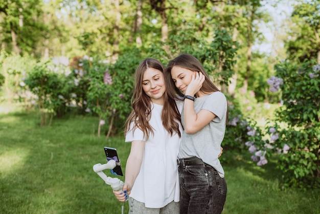 Lächelnde hübsche junge Influencer-Bloggerinnen filmen oder nehmen Videos mit ihrem Handy auf einem Stabilisator in einem sonnigen grünen Park draußen auf. Blogging-Konzept. Weicher selektiver Fokus.