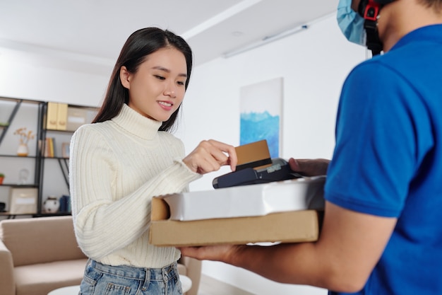 Lächelnde hübsche junge Vietnamesin, die Kreditkarte durchzieht, um den Kurier für die Pizzalieferung zu bezahlen?