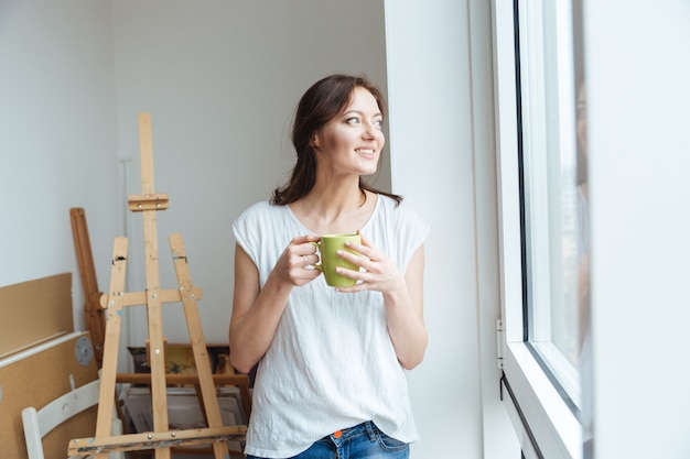 Lächelnde hübsche künstlerin trinkt kaffee am fenster in der werkstatt