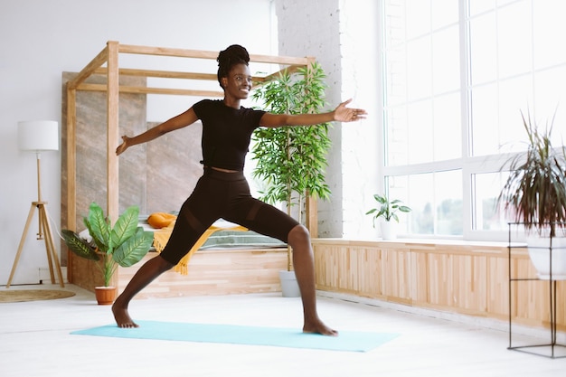 Lächelnde interrassische Frau macht zu Hause Yoga auf der Matte Halten Sie die Hände gerade über dem Kopf in Virabhadrasana Warrior Two