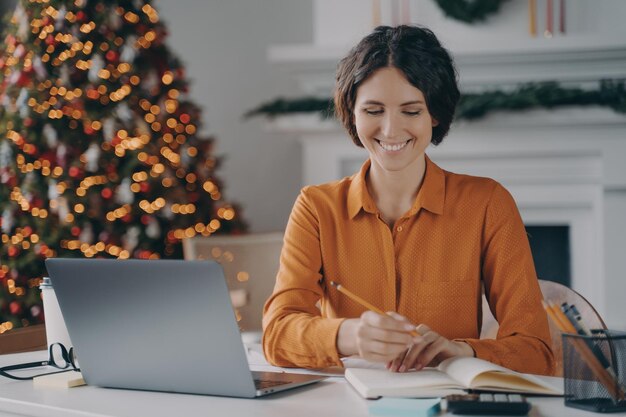 Lächelnde italienische Frau, die während der Weihnachtsferien Notizen mit Laptop zu Hause macht