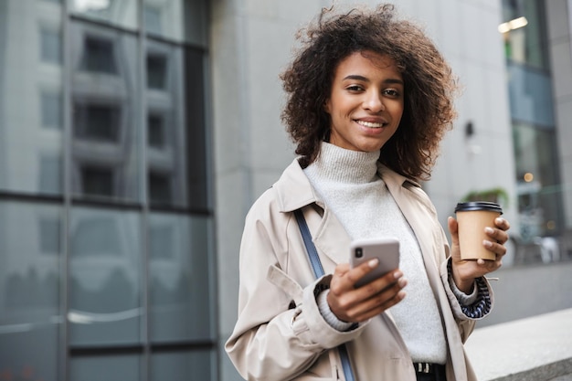 Lächelnde junge Afrikanerin im Herbstmantel, die im Freien spazieren geht, mit Handy