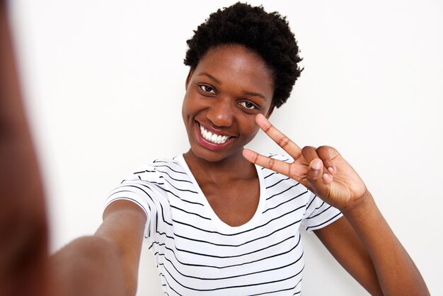 Lächelnde junge afrikanische Frau, die Friedenszeichen selfie gegen weißen Hintergrund bildet