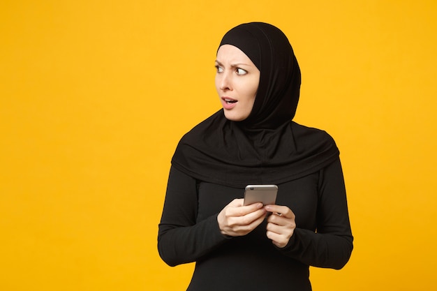 Lächelnde junge arabische muslimische Frau in schwarzer Hijab-Kleidung hält in den Händen Handy und plaudert isoliert auf gelbem Wandporträt. Menschen religiöses Lifestyle-Konzept. .