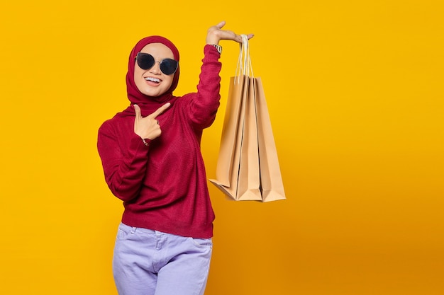 Lächelnde junge Asiatin, die mit dem Finger auf Einkaufstüten auf gelbem Hintergrund zeigt