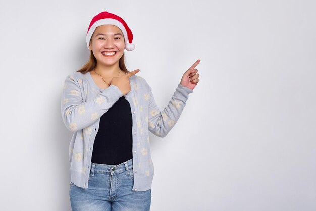 Lächelnde junge Asiatin in einem Weihnachtsmann-Hut, die mit dem Finger auf den leeren Kopienraum zeigt, der über weißem Hintergrund isoliert ist