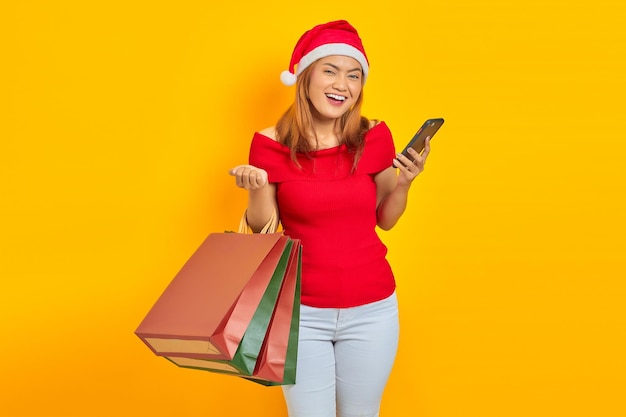 Lächelnde junge Asiatin mit Einkaufstüten und Handy auf gelbem Hintergrund isoliert