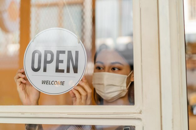 Lächelnde junge asiatische Besitzerin im Einzelhandel, Café-Frau, die das Schild zum Öffnen mit Gesichtsmaske umdreht, sich vor einer Pandemie des Coronavirus schützt, das Geschäft nach der Schließung der Quarantäne in Covid bis zur neuen Normalität wiedereröffnet.