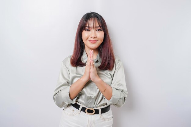 Lächelnde junge asiatische Frau, die das salbeigrüne Hemd trägt, das den traditionellen Gruß lokalisiert über weißem Hintergrund gestikuliert