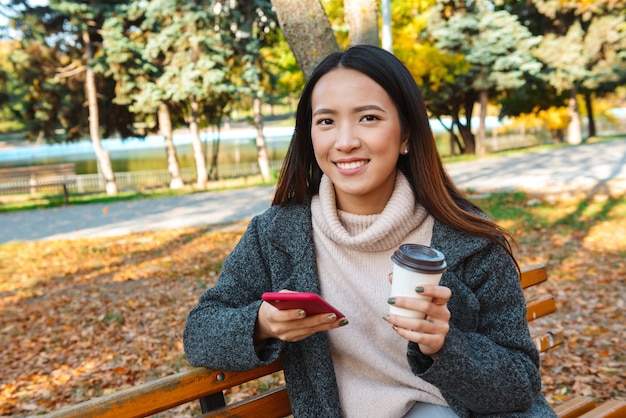 Lächelnde junge asiatische Frau, die Mantel sitzt, sitzt auf einer Bank im Park, mit Handy