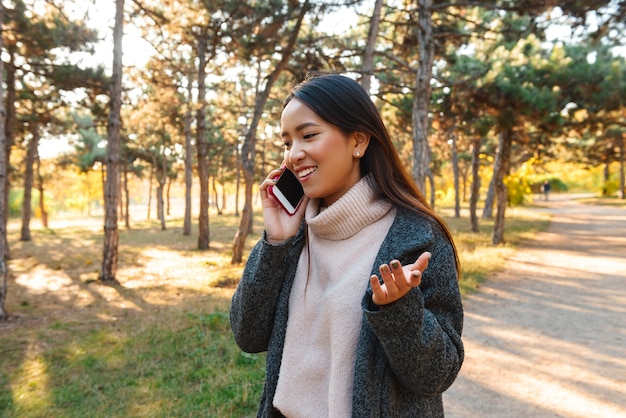 Lächelnde junge asiatische Frau, die Mantel trägt, der draußen im Park geht und auf Handy spricht