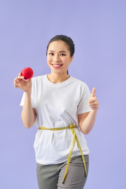 Lächelnde junge asiatische Frau, die Maßband um ihre Taille hält und roten Apfel über violettem Hintergrund isst