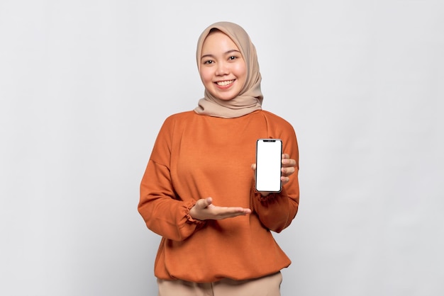 Lächelnde junge asiatische Frau im orangefarbenen Hemd, das den leeren Bildschirm des Handys zeigt und die App isoliert über weißem Hintergrund empfiehlt