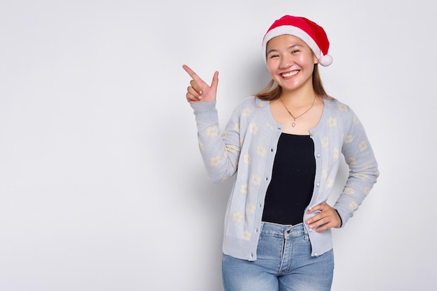 Lächelnde junge asiatische Frau in einem Weihnachtsmann-Hut, der mit dem Finger auf den Kopienraum zeigt, der über weißem Hintergrund isoliert ist