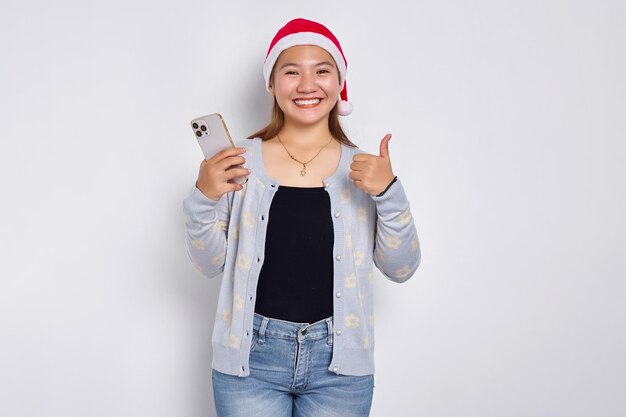 Lächelnde junge asiatische Frau in einer Weihnachtsmütze, die Smartphone verwendet und den Daumen lokalisiert über weißem Hintergrund zeigt