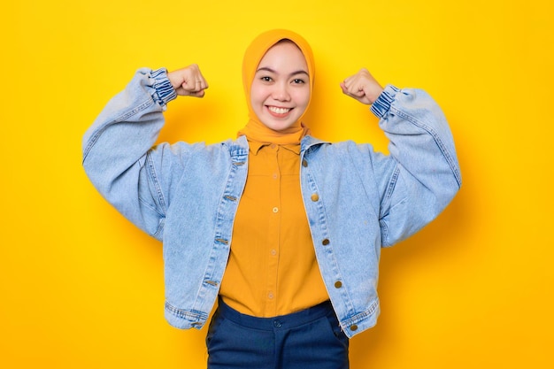 Lächelnde junge asiatische Frau in Jeansjacke hebt ihre Arme und zeigt Muskeln isoliert auf gelbem Hintergrund