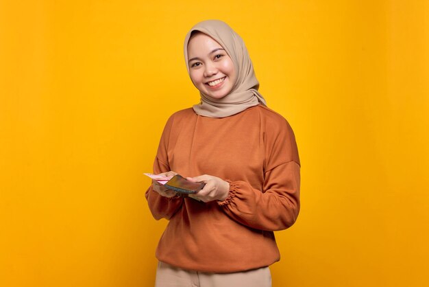 Lächelnde junge asiatische Frau in orangefarbenem Hemd, die eine Brieftasche voller Geldscheine zeigt, die über gelbem Hintergrund isoliert sind