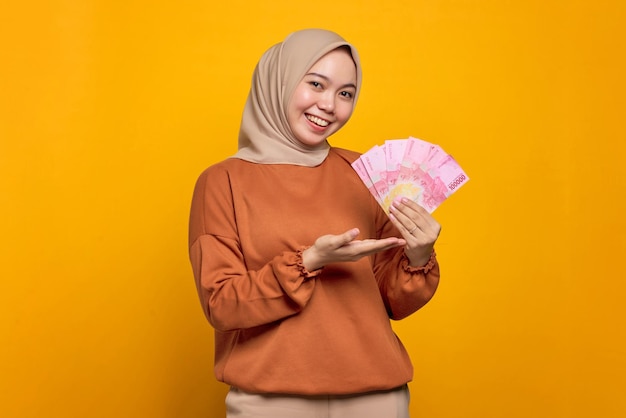 Lächelnde junge asiatische Frau in orangefarbenem Hemd, die Geldscheine einzeln auf gelbem Hintergrund zeigt
