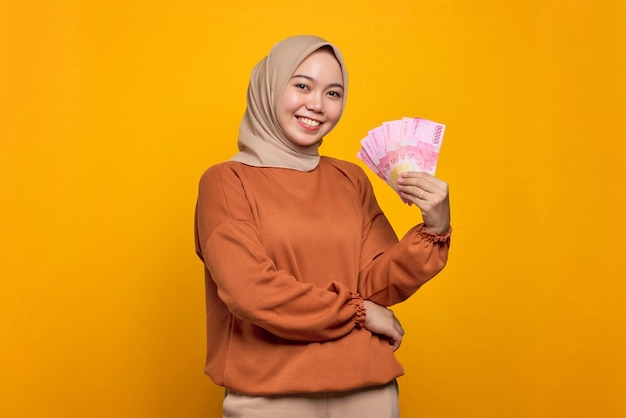 Lächelnde junge asiatische Frau in orangefarbenem Hemd mit Geldbanknoten isoliert auf gelbem Hintergrund