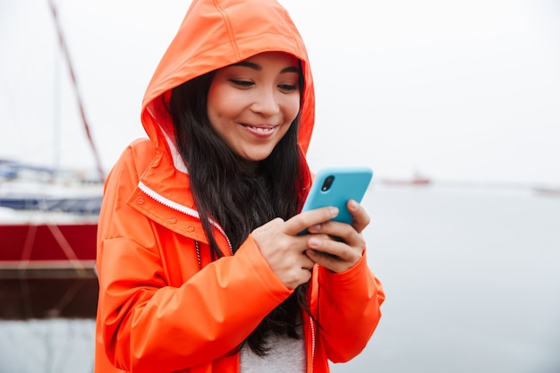 Lächelnde junge asiatische Frau mit Regenmantel, die Zeit im Freien verbringt, an der Küste spazieren, mit Handy