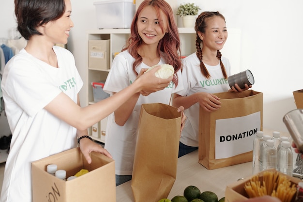 Lächelnde junge asiatische Frauen, die Konserven, Obst, Gemüse und Wasser in Papierverpackungen verpacken, um in einem wohltätigen Zentrum zu spenden