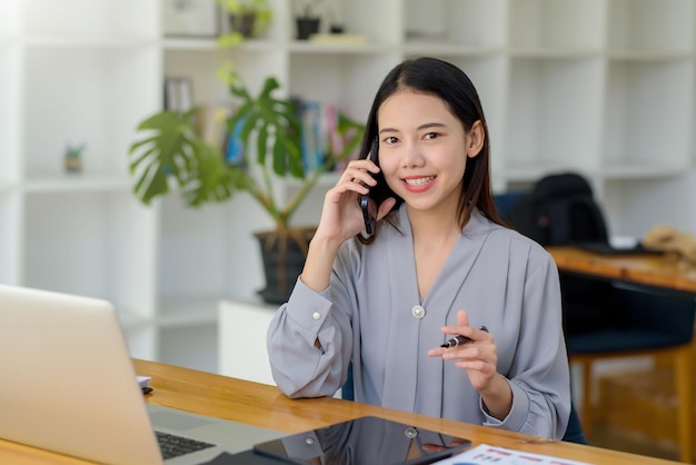 Lächelnde junge asiatische Geschäftsfrau, die am Telefon mit Laptop-Computer auf dem Schreibtisch im Büro spricht