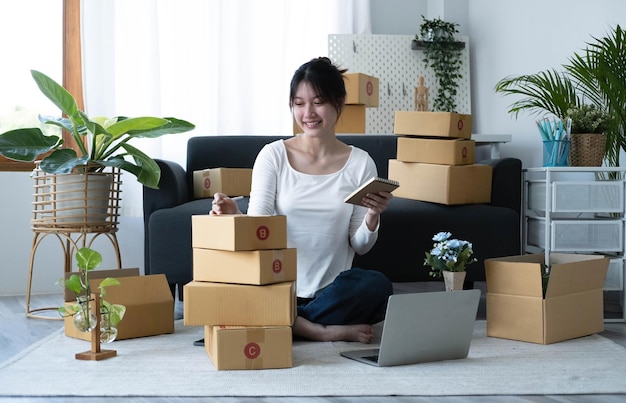 Lächelnde junge asiatische Geschäftsinhaberin bereiten Paketboxen und Online-Bestellungen von Produkten für die Lieferung an Kunden auf Laptop-Computern vor Online-Shopping-Konzept