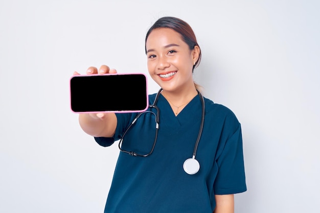 Lächelnde junge asiatische Krankenschwester, die in einer blauen Uniform arbeitet und ein Smartphone mit leerem Bildschirm zeigt, der auf weißem Hintergrund isoliert ist. Konzept der Gesundheitsmedizin