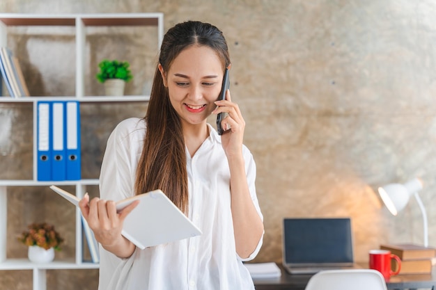 Lächelnde junge asiatische Managerin, die aus der Ferne arbeitet und Papier hält und Finanzdokumente liest Gespräch über ein Handy, das zu Hause an einem Schreibtisch steht